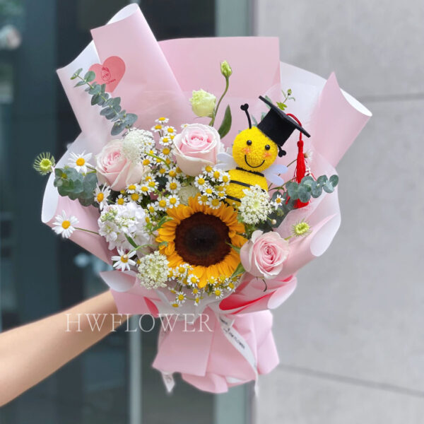 Pink graduation bouquet featuring a sunflower, pink roses, white daisies, and a cute bee plush in pink wrapping.