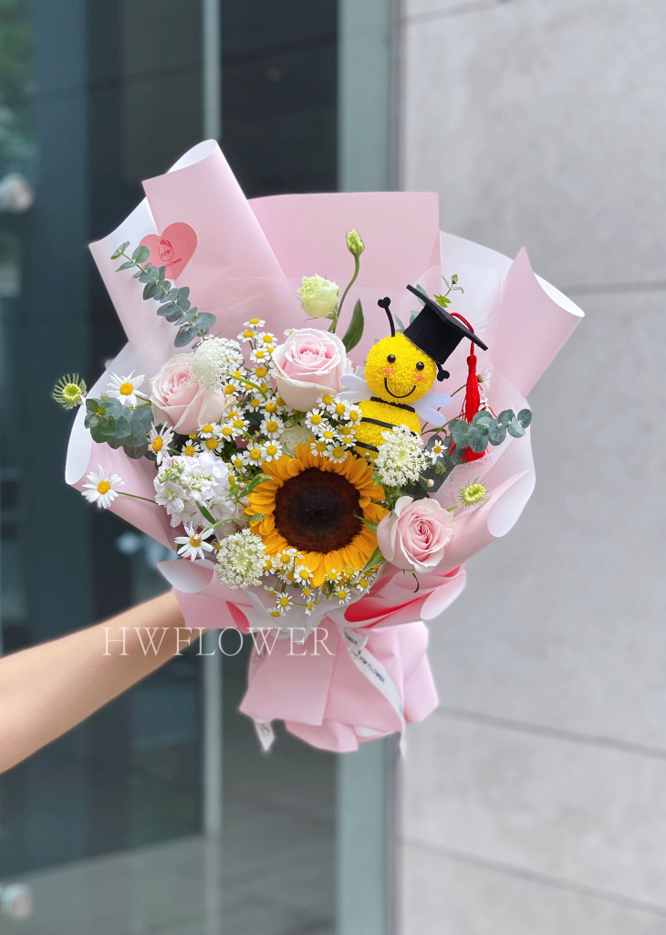 Pink graduation bouquet featuring a sunflower, pink roses, white daisies, and a cute bee plush in pink wrapping.