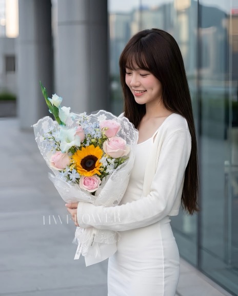 A woman in a white outfit holding a lace-wrapped bouquet of pink roses, a sunflower and pale blue flowers.|一位穿著白色服裝的女生手持蕾絲布包花花束,花束配有粉紅玫瑰、向日葵及淺藍色花朵。