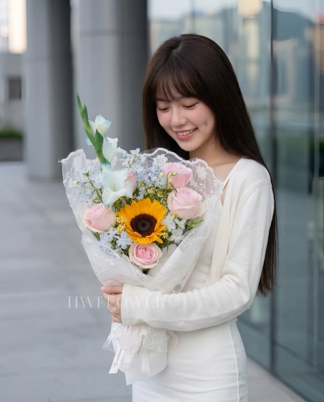 A woman in a white outfit holding a bouquet of pink roses, a sunflower and pale blue flowers wrapped in white lace fabric.